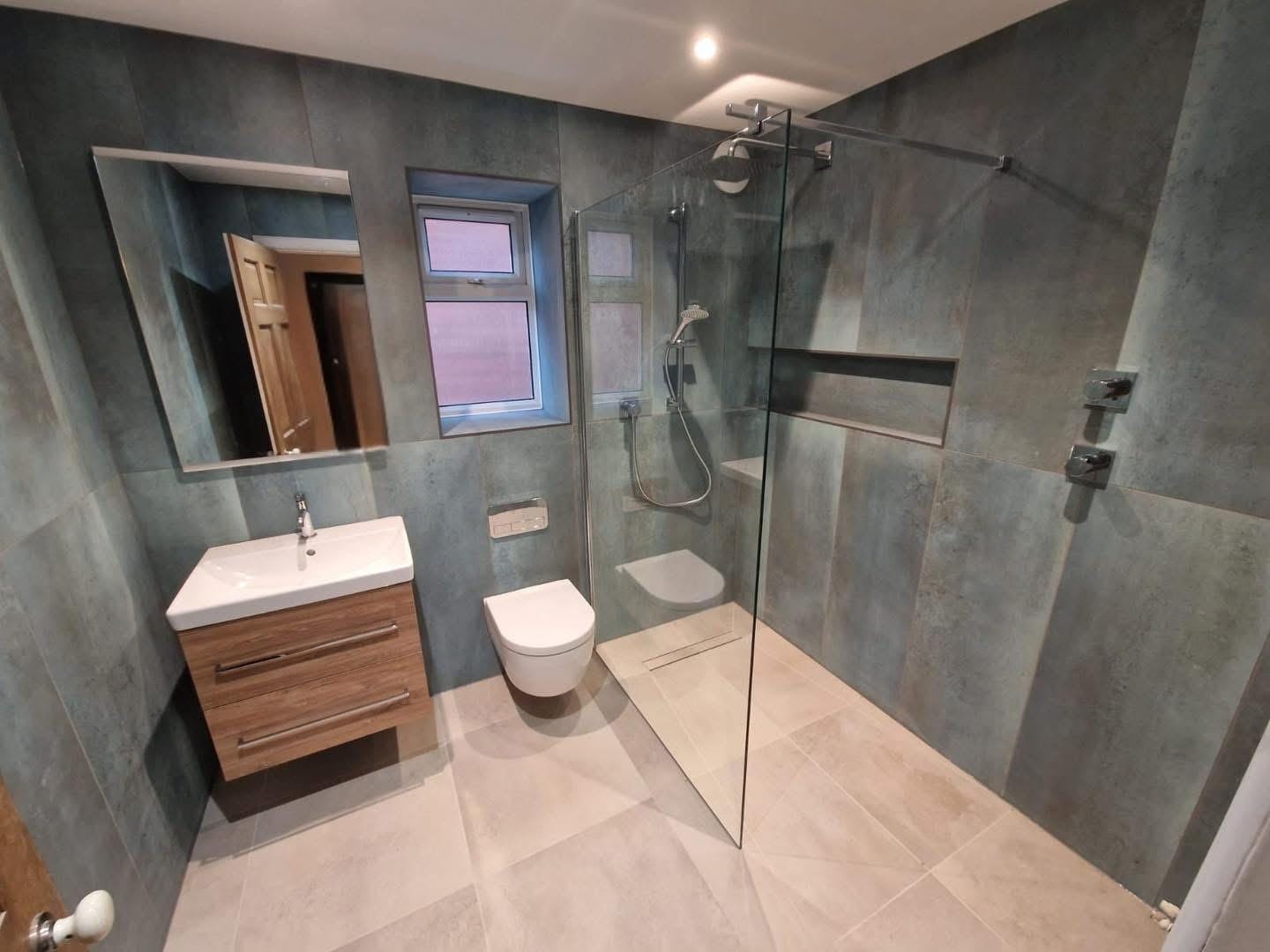 The finished wet room, tiled in blue grey stone effect with a wall hung wood vanity, a wall hung WC and a level access shower behind a glass screen