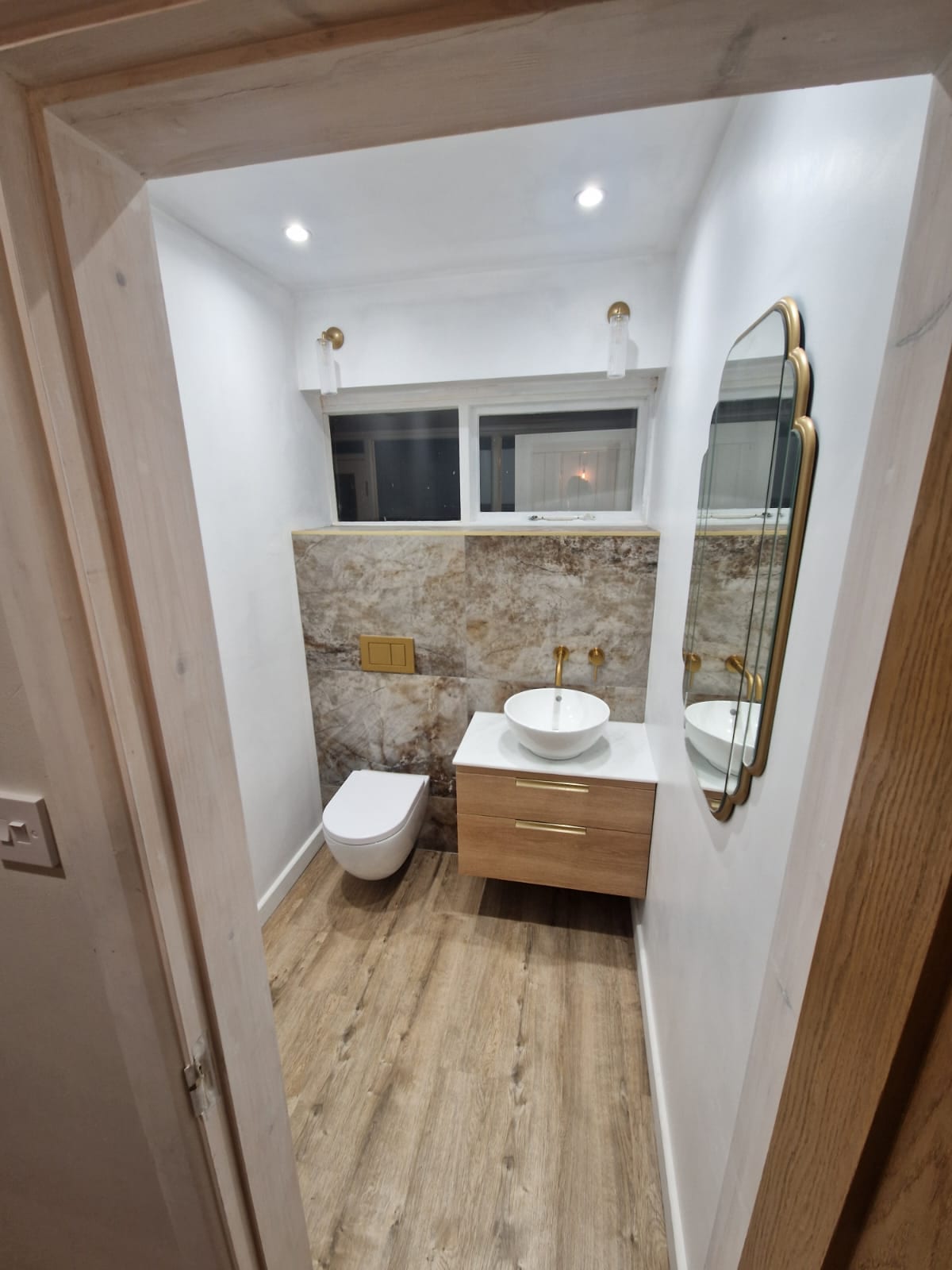 The finished cloakroom, with a wall hung WC and a wood vanity with a marble top, against grey brown marble effect tiling and brass fittings